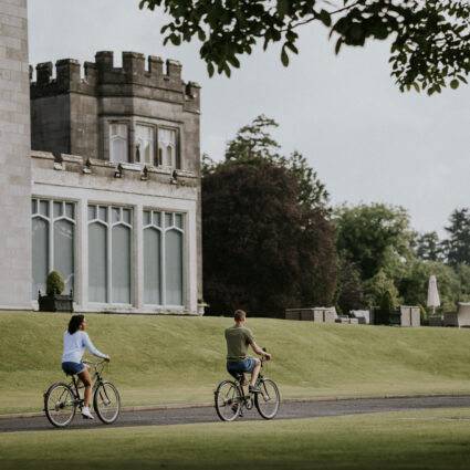 Guests enjoying a leisurely cycle on scenic hotel grounds with lush gardens, near a charming historic building.