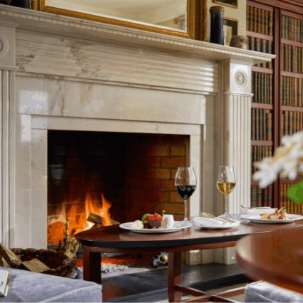 Guests enjoy fine dining with exquisite desserts and wine by a roaring fireplace in Dromoland Castle's opulent library, surrounded by antique books.