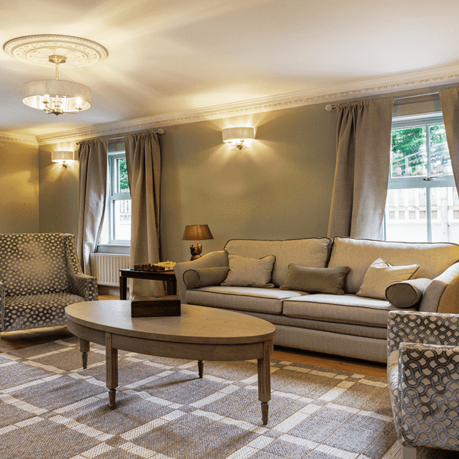 Spacious modern hotel suite with elegant furnishings, natural light, cosy sofa, armchairs, desk, and warm tones.