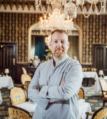 Chef in elegant dining room with chandeliers, ornate decor, and plush seating at luxury hotel.