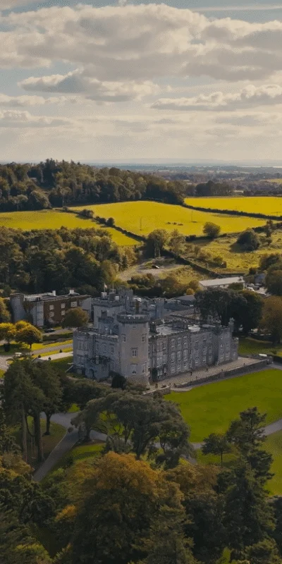 Aerial view of luxurious countryside hotel with castle architecture, lush gardens, scenic landscape, and serene lake.