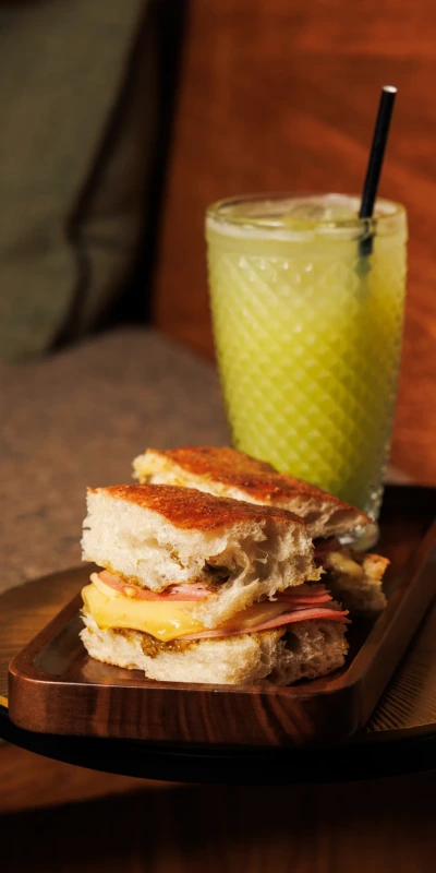 A gourmet sandwich with layers of cheese and ham on a wooden tray next to a frothy green beverage, set against a rustic backdrop. Dromoland, Newmarket on Fergus, Co. Clare, Ireland.